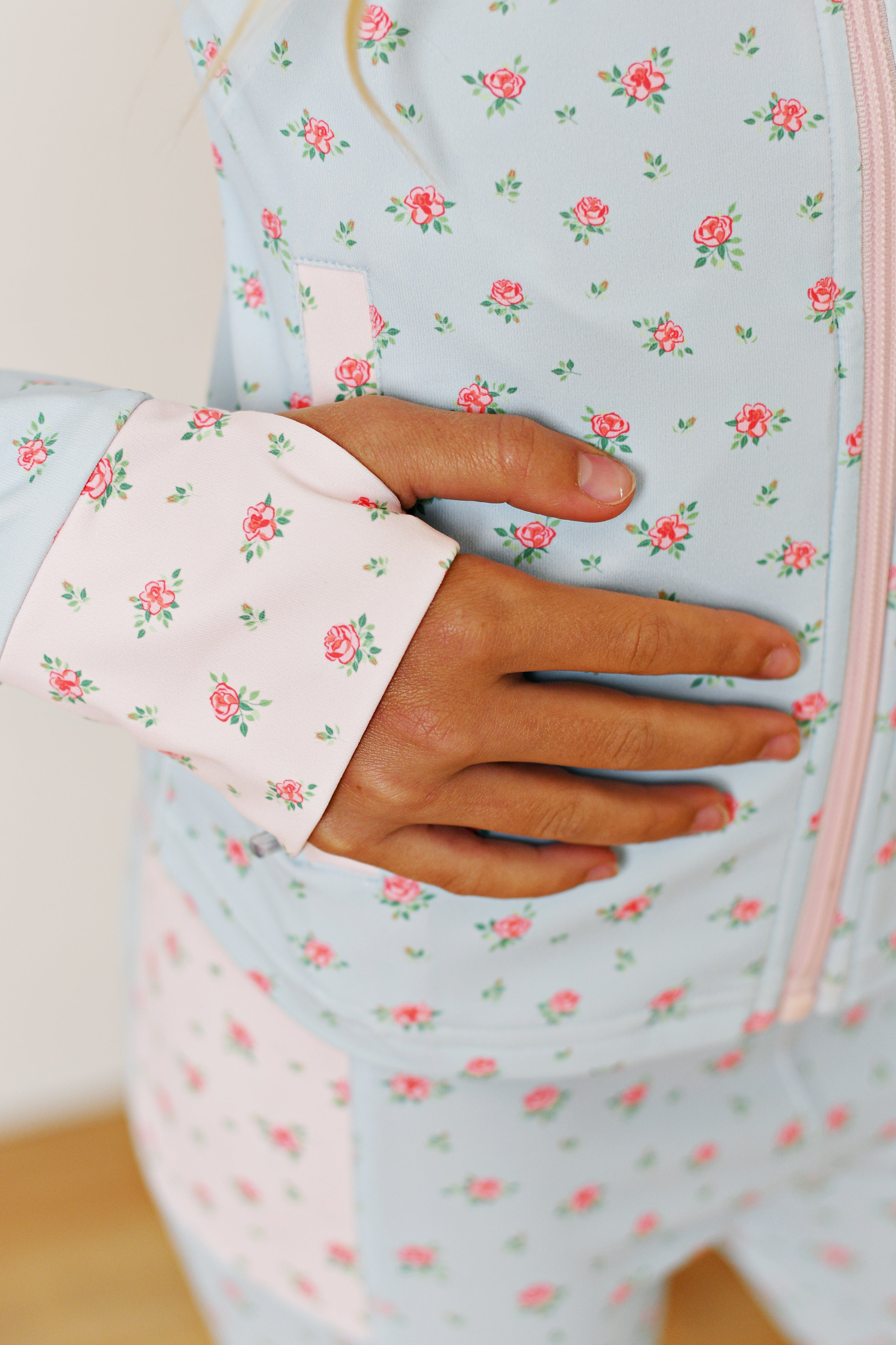 A close-up of a person wearing a light blue floral zip-up jacket with pink and green rose patterns, showing a hand resting on the front pocket.