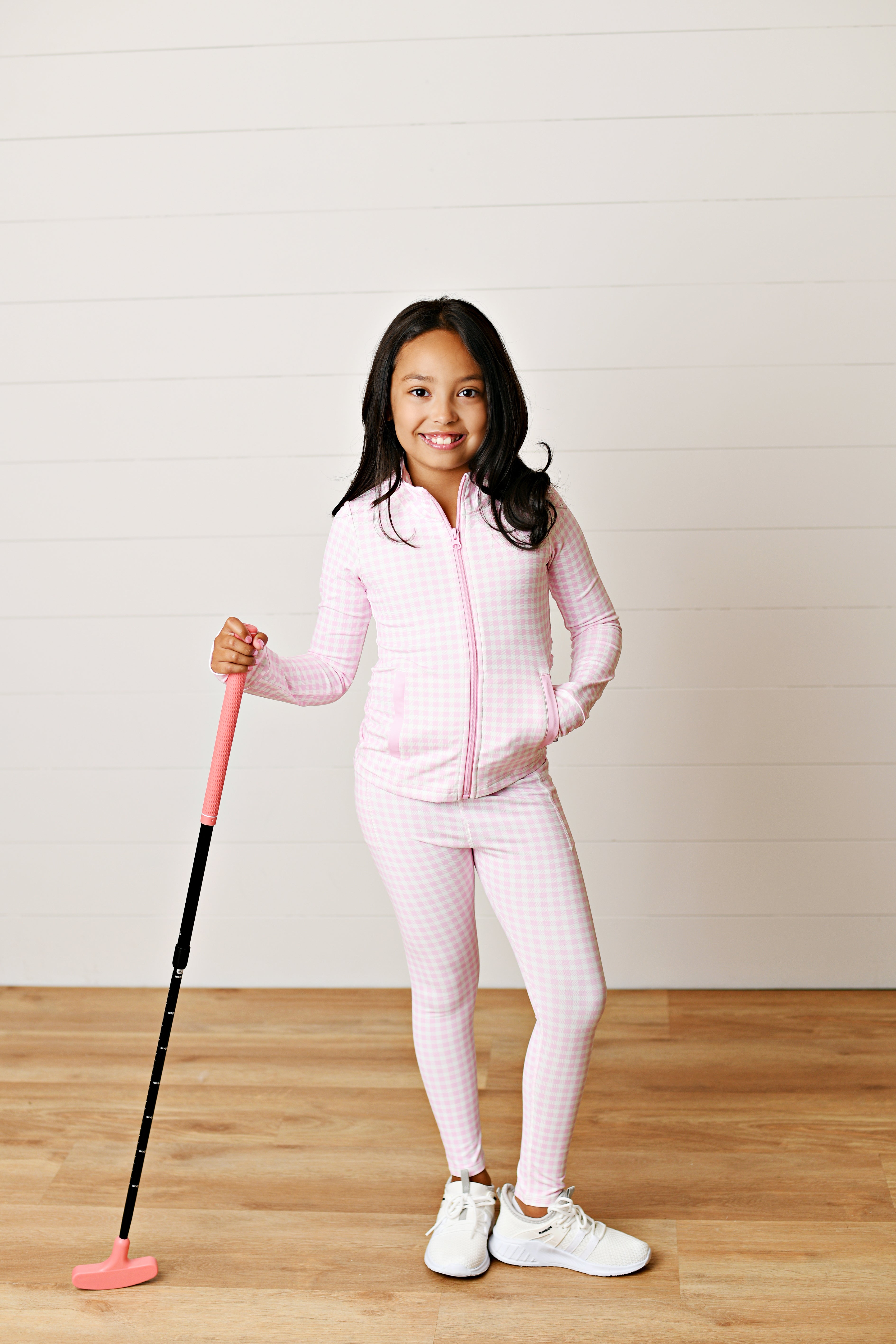 A young girl stands on a wooden floor against a white paneled wall, wearing a light pink and white gingham zip-up jacket and matching leggings, holding a pink and black golf club.