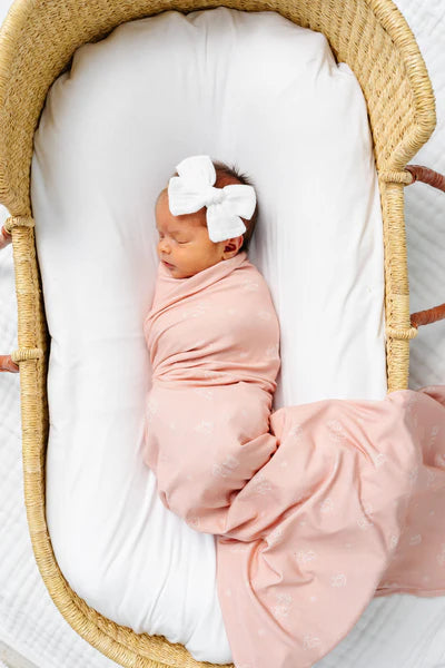 Newborn baby wrapped in pink swaddle with a white bow in a wooden crib.