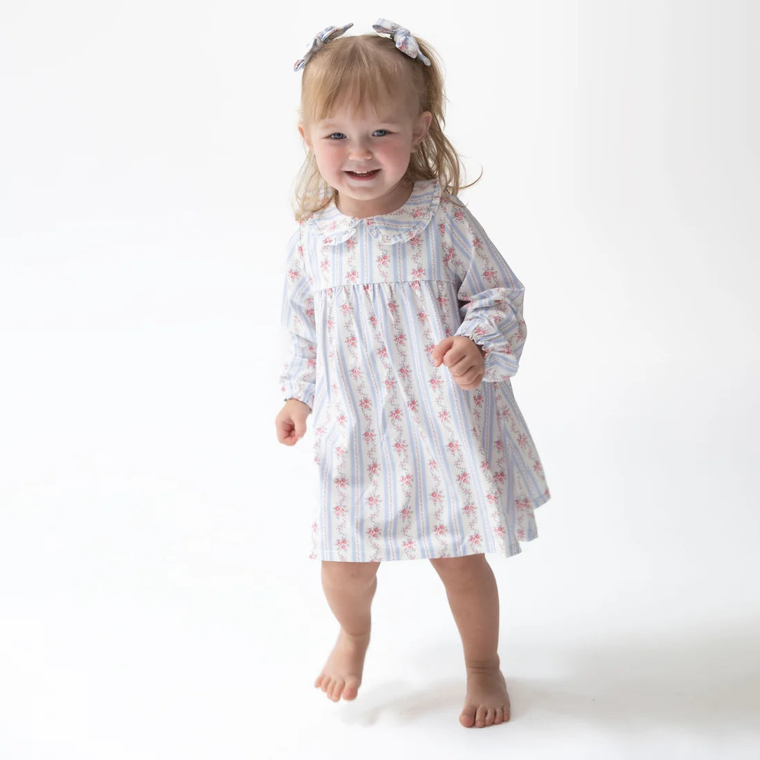 A toddler girl wearing a long-sleeve dress with a blue and white vertical stripe pattern and a small pink floral print, standing barefoot against a plain white studio background.