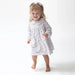 A toddler girl wearing a long-sleeve dress with a blue and white vertical stripe pattern and a small pink floral print, standing barefoot against a plain white studio background.