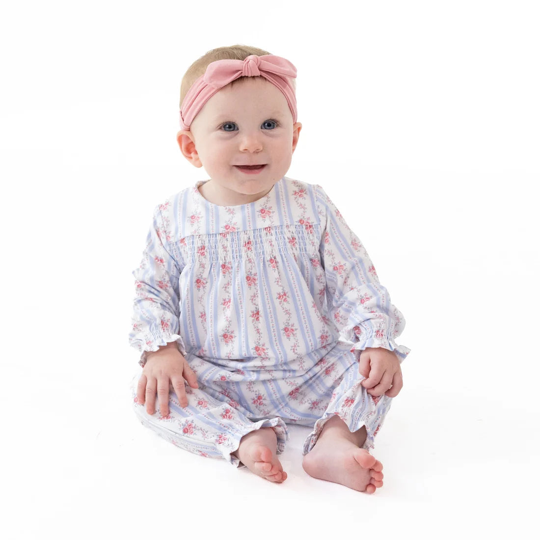 A baby girl sitting on a white studio background, wearing a long-sleeve smocked romper in a light blue and white floral pattern with a pink bow headband.