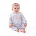 A baby girl sitting on a white studio background, wearing a long-sleeve smocked romper in a light blue and white floral pattern with a pink bow headband.