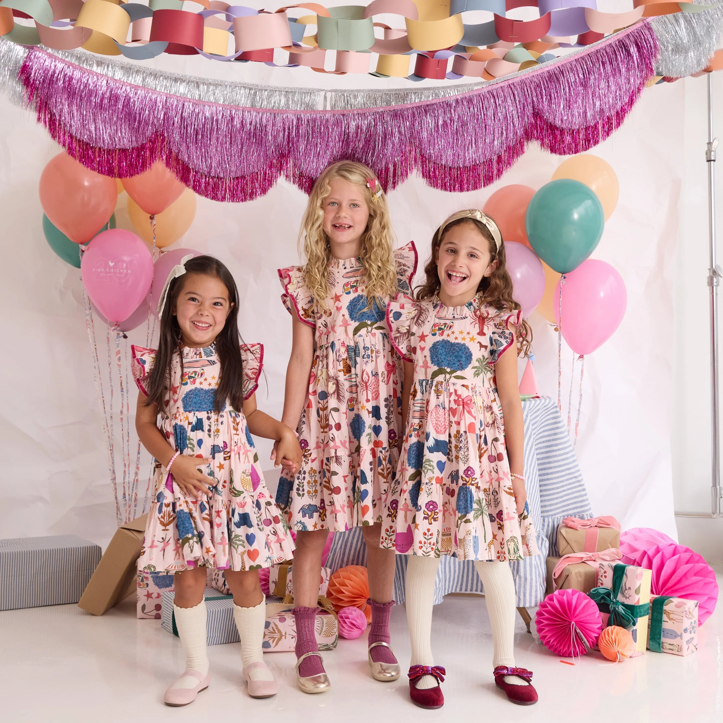 Three young girls wearing matching floral print dresses stand together in a decorated studio setting with balloons, tinsel garlands, and party decorations. The dresses feature a multicolor pattern of blue, pink, orange, and green on a light background.