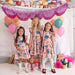 Three young girls wearing matching floral print dresses stand together in a decorated studio setting with balloons, tinsel garlands, and party decorations. The dresses feature a multicolor pattern of blue, pink, orange, and green on a light background.