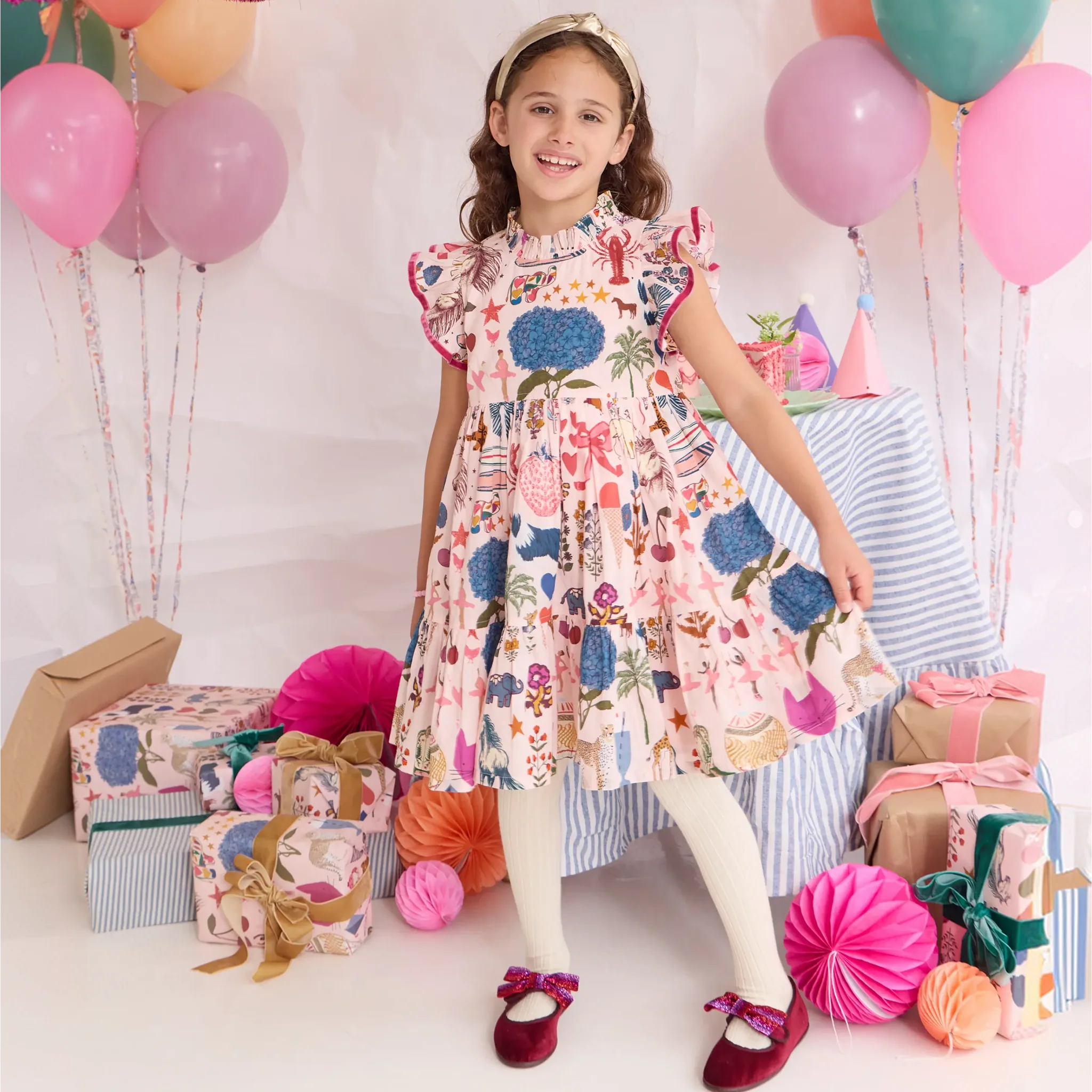A young girl stands in a decorated party setting wearing a short-sleeve ruffled dress with a colorful floral and whimsical print. She is surrounded by balloons, wrapped gifts, paper decorations, and a striped tablecloth.