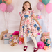 A young girl stands in a decorated party setting wearing a short-sleeve ruffled dress with a colorful floral and whimsical print. She is surrounded by balloons, wrapped gifts, paper decorations, and a striped tablecloth.