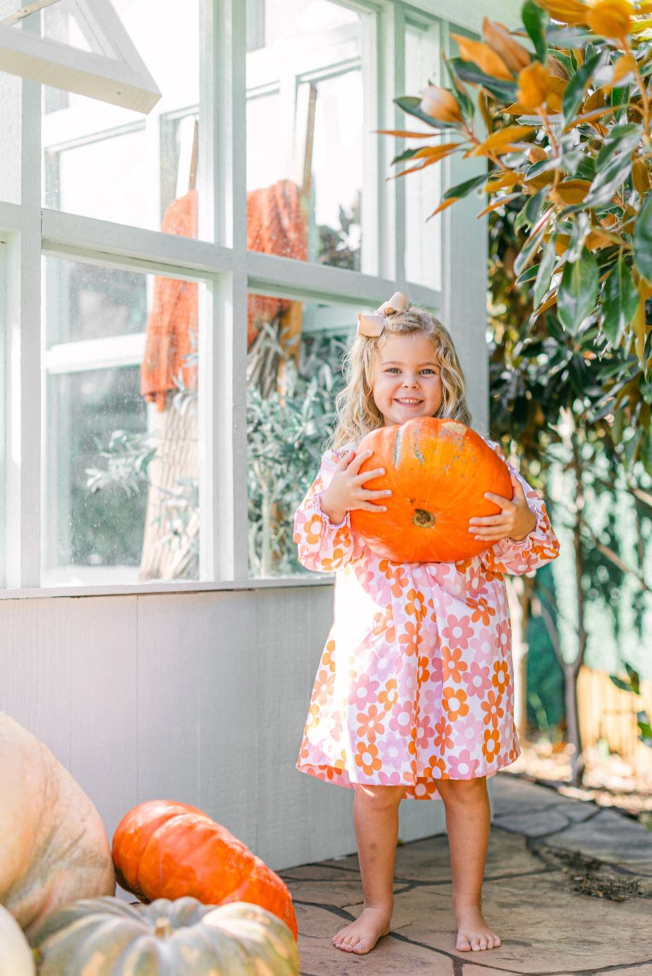Poppy Kids pink and orange floral girls dress with long sleeves modeled by a child holding a pumpkin in a bright autumn setting.
