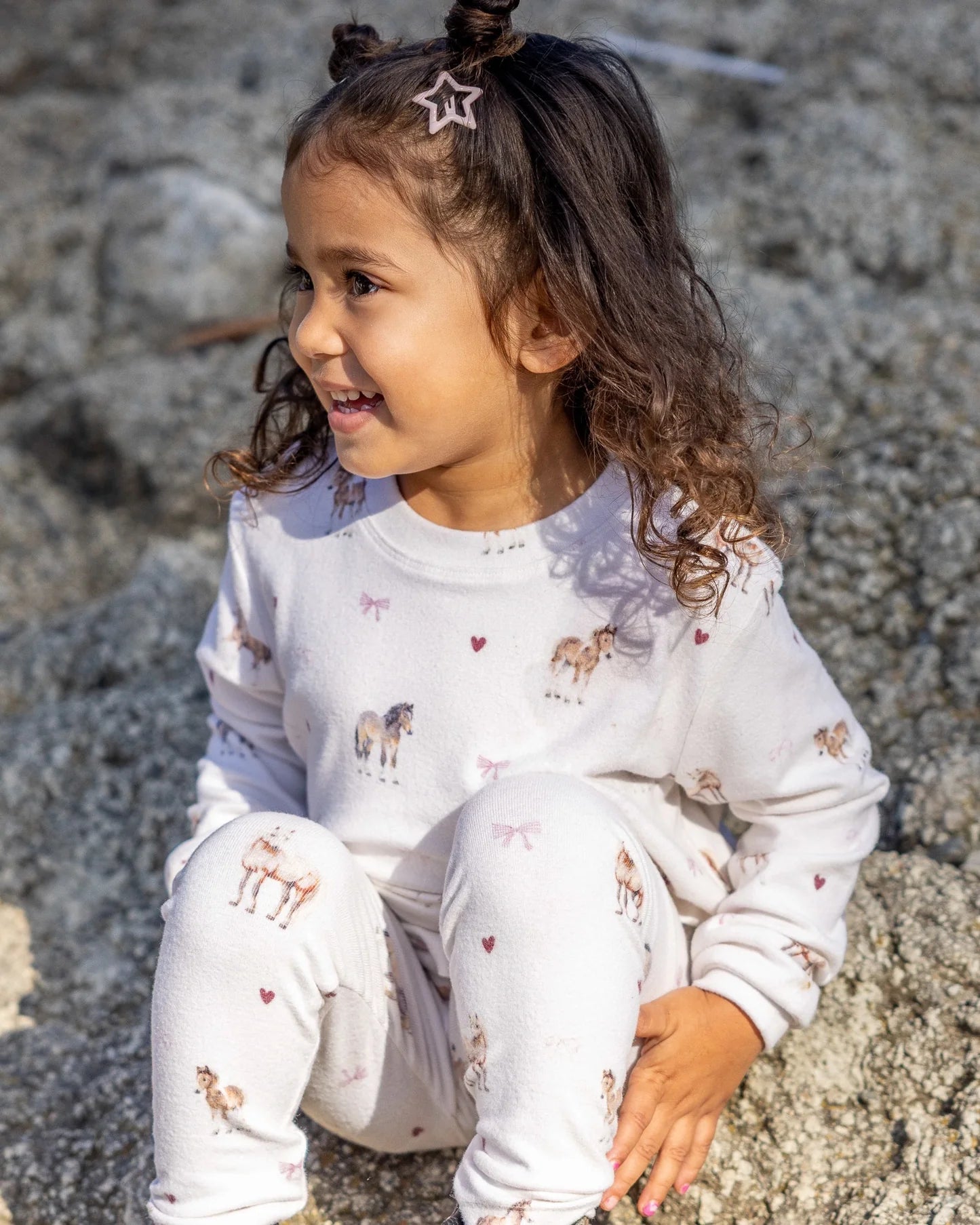 A young girl with curly brown hair sits on a rock outdoors, wearing a white long-sleeve sweatsuit with a cute print of horses and small pink hearts.