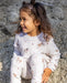 A young girl with curly brown hair sits on a rock outdoors, wearing a white long-sleeve sweatsuit with a cute print of horses and small pink hearts.