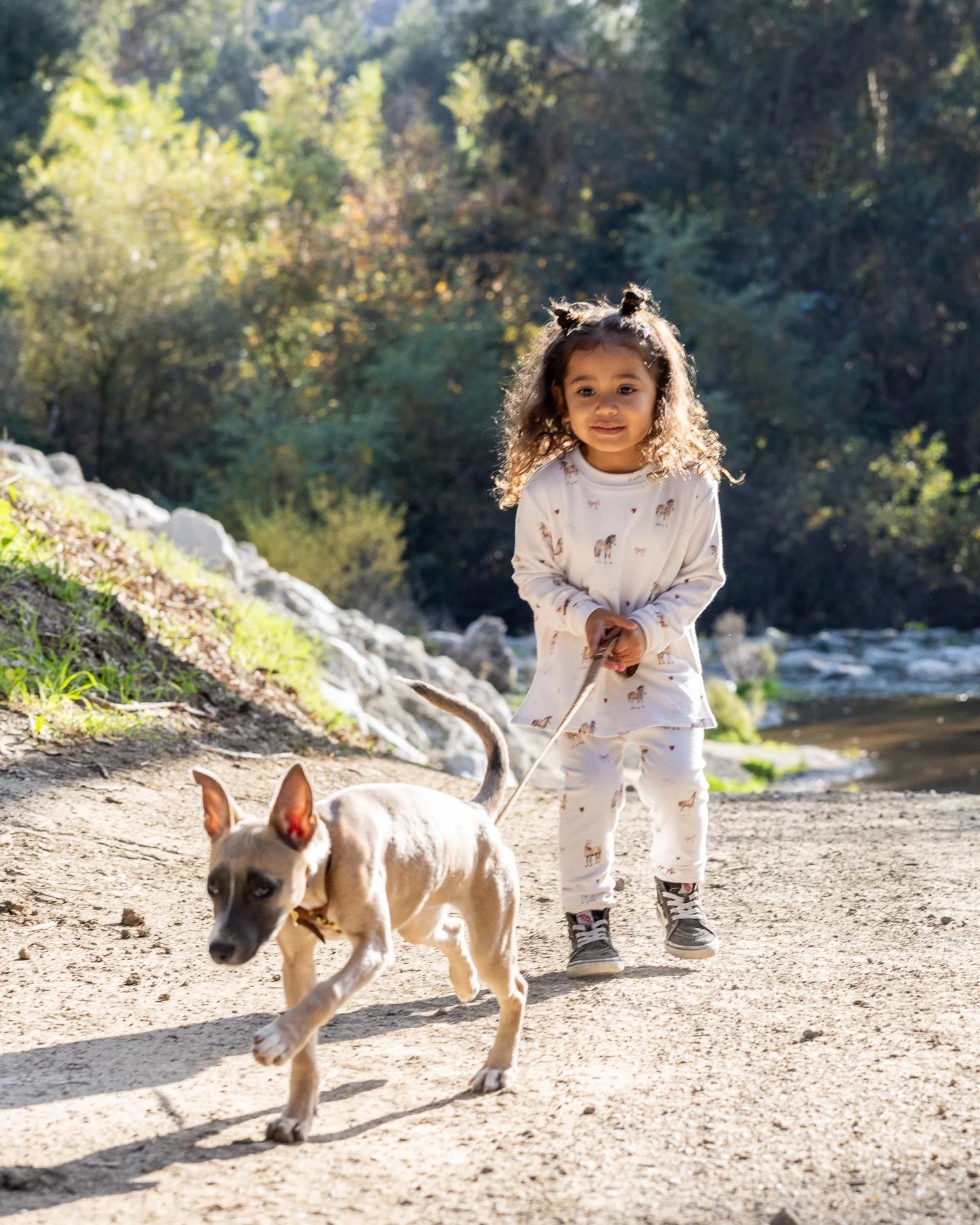 A young child wearing a matching light-colored sweatsuit walks outdoors on a dirt path with a small dog, with trees and rocks in the background.