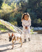A young child wearing a matching light-colored sweatsuit walks outdoors on a dirt path with a small dog, with trees and rocks in the background.