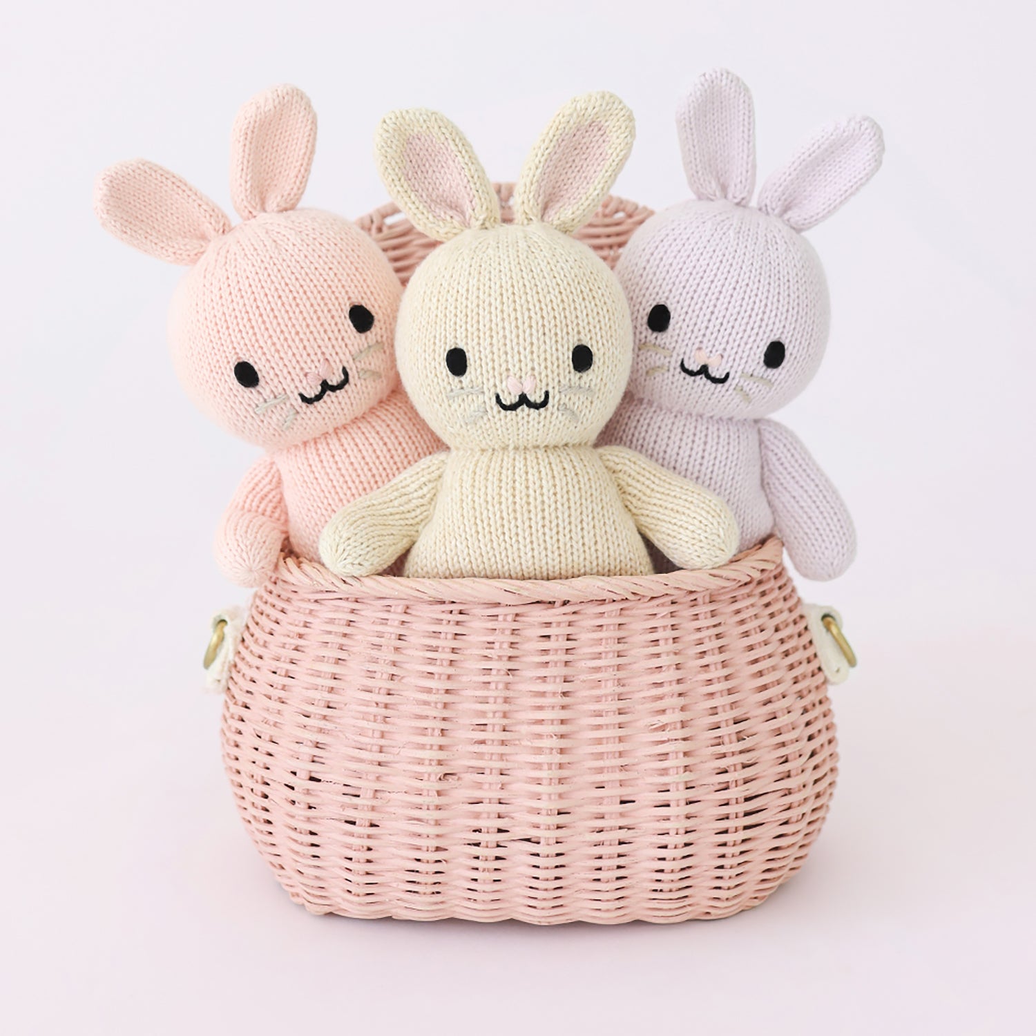 Three knitted bunny toys in a pink basket on a light pink background