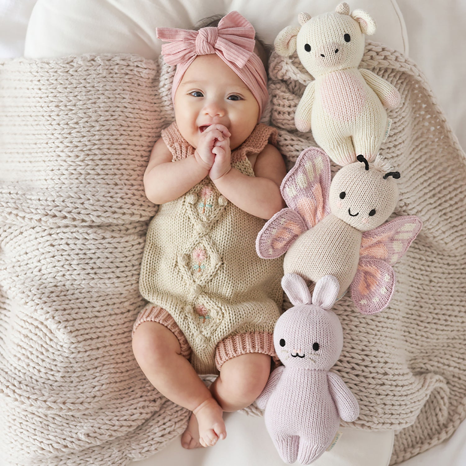 Baby lying on a soft surface with knitted toys, wearing a pink headband and beige outfit.