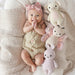 Baby lying on a soft surface with knitted toys, wearing a pink headband and beige outfit.