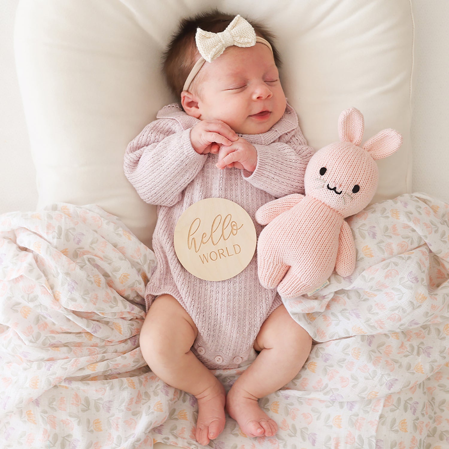 Newborn baby in a pink onesie with a 'hello world' tag, holding a pink knitted bunny toy.