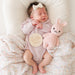 Newborn baby in a pink onesie with a 'hello world' tag, holding a pink knitted bunny toy.