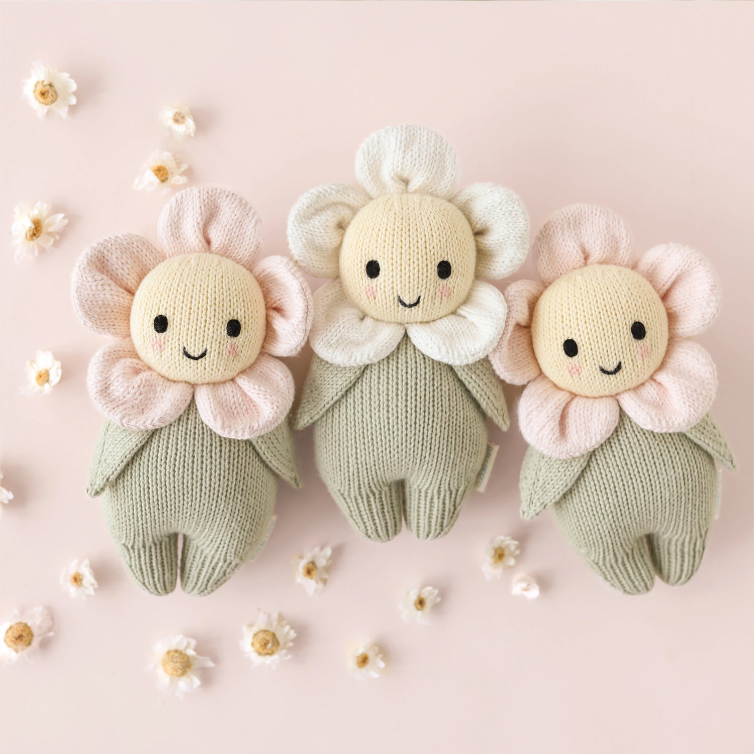 Three knitted flower dolls on a pink background with small flowers around them.