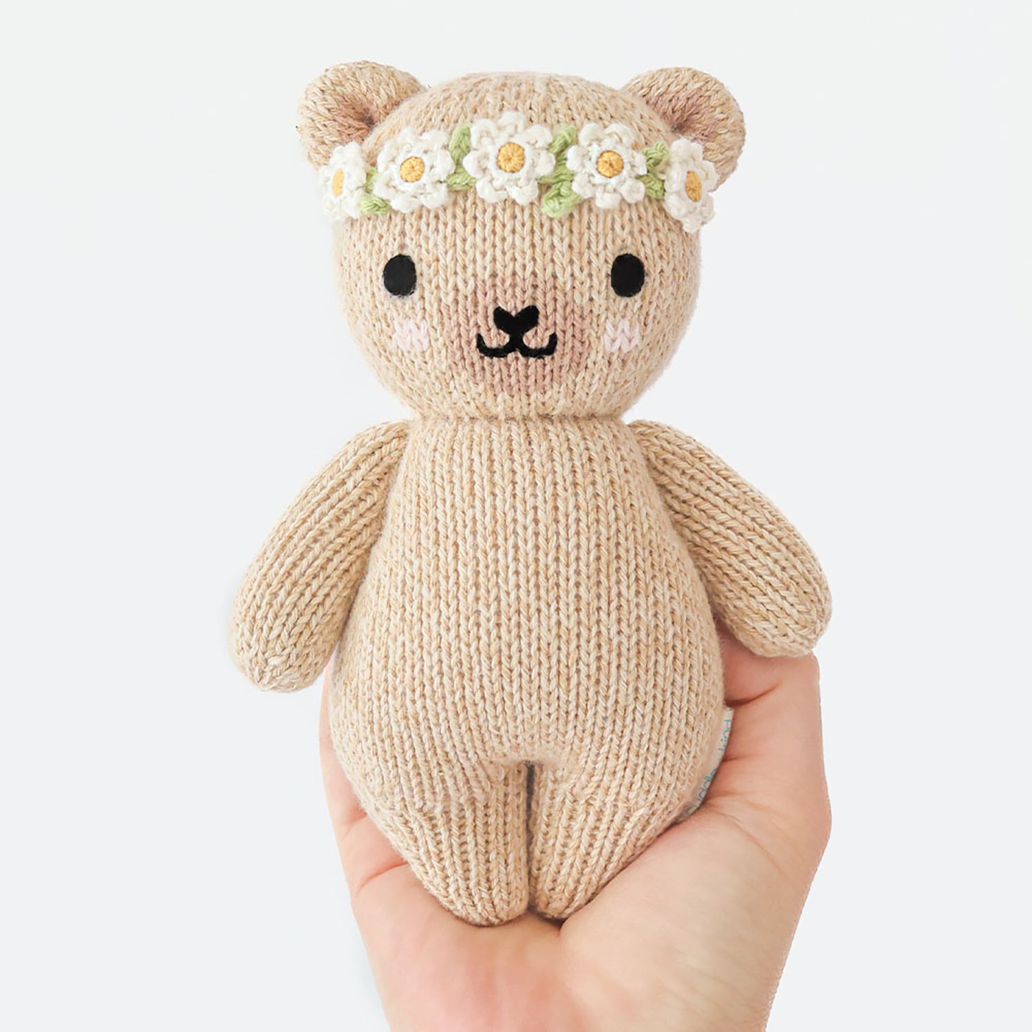 Knitted teddy bear with a flower crown held in a hand against a white background