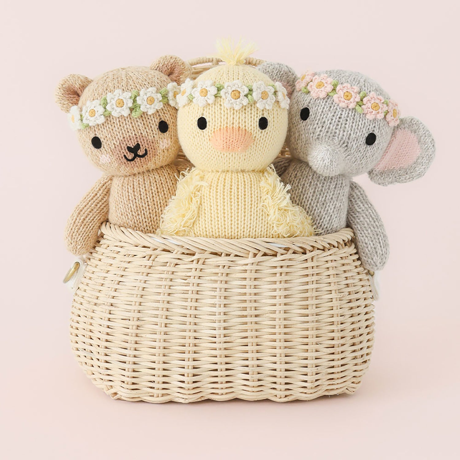 Three knitted toys, resembling a bear, a lamb, and a mouse, sitting in a woven basket on a light pink background.