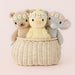 Three knitted toys, resembling a bear, a lamb, and a mouse, sitting in a woven basket on a light pink background.