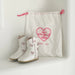 White children's cowboy boots with pink accents next to a 'Tiny Cowgirl Club' drawstring bag on a light background