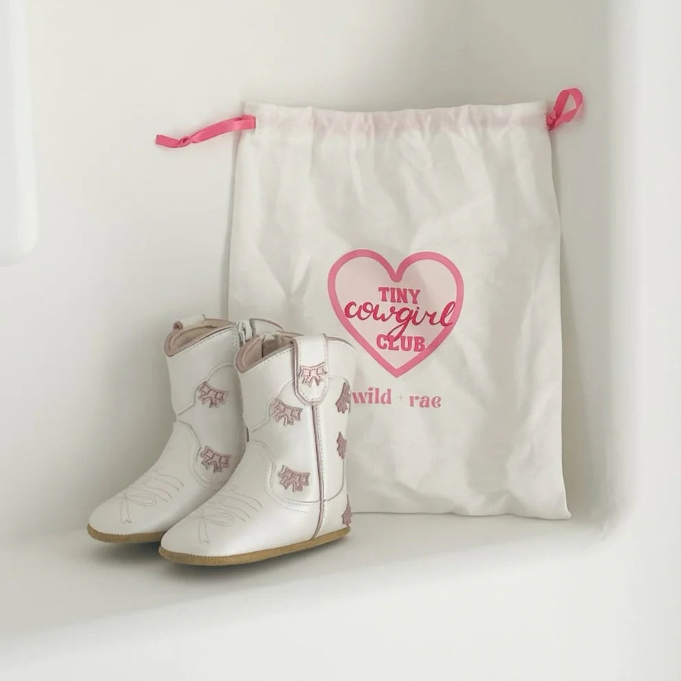 White children's cowboy boots with pink accents next to a 'Tiny Cowgirl Club' drawstring bag on a light background