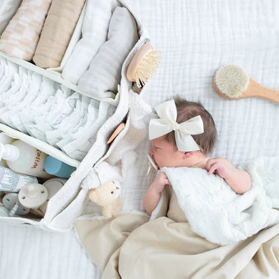 A white textured tote bag with dual top handles, laying next to a newborn baby. A small beige teddy bear keychain hangs from the handle.