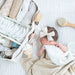 A white textured tote bag with dual top handles, laying next to a newborn baby. A small beige teddy bear keychain hangs from the handle.
