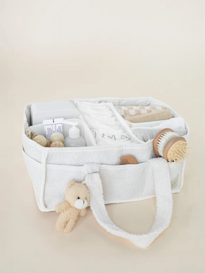 A light cream fabric baby toiletry bag filled with baby items, including a folded towel, a small teddy bear, a pump bottle, and a wooden brush, set against a solid beige background.
