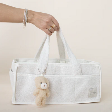 A white textured tote bag with dual top handles, held by a person's hand. A small beige teddy bear keychain hangs from the handle.