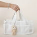 A white textured tote bag with dual top handles, held by a person's hand. A small beige teddy bear keychain hangs from the handle.