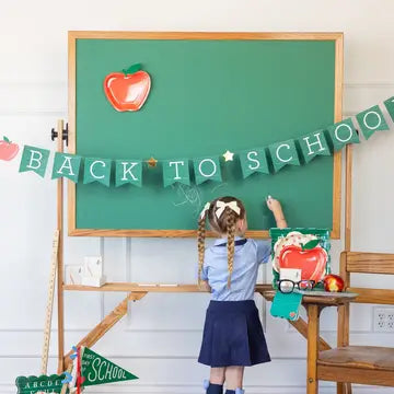 Child in school uniform standing next to a green chalkboard with 'Back to School' decorations.