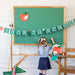 Child in school uniform standing next to a green chalkboard with 'Back to School' decorations.