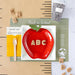 Red apple-shaped plate with 'ABC' letters on a back-to-school themed background with a black and white composition cup