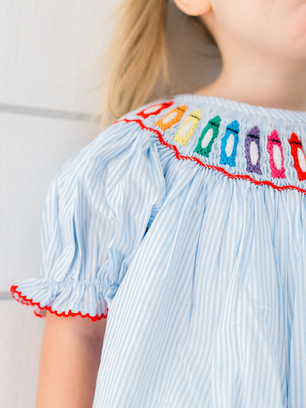 Close-up of a child wearing a light blue and white vertically striped smocked dress with colorful crayon embroidered details around the neckline and red scalloped trim.