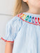 Close-up of a child wearing a light blue and white vertically striped smocked dress with colorful crayon embroidered details around the neckline and red scalloped trim.