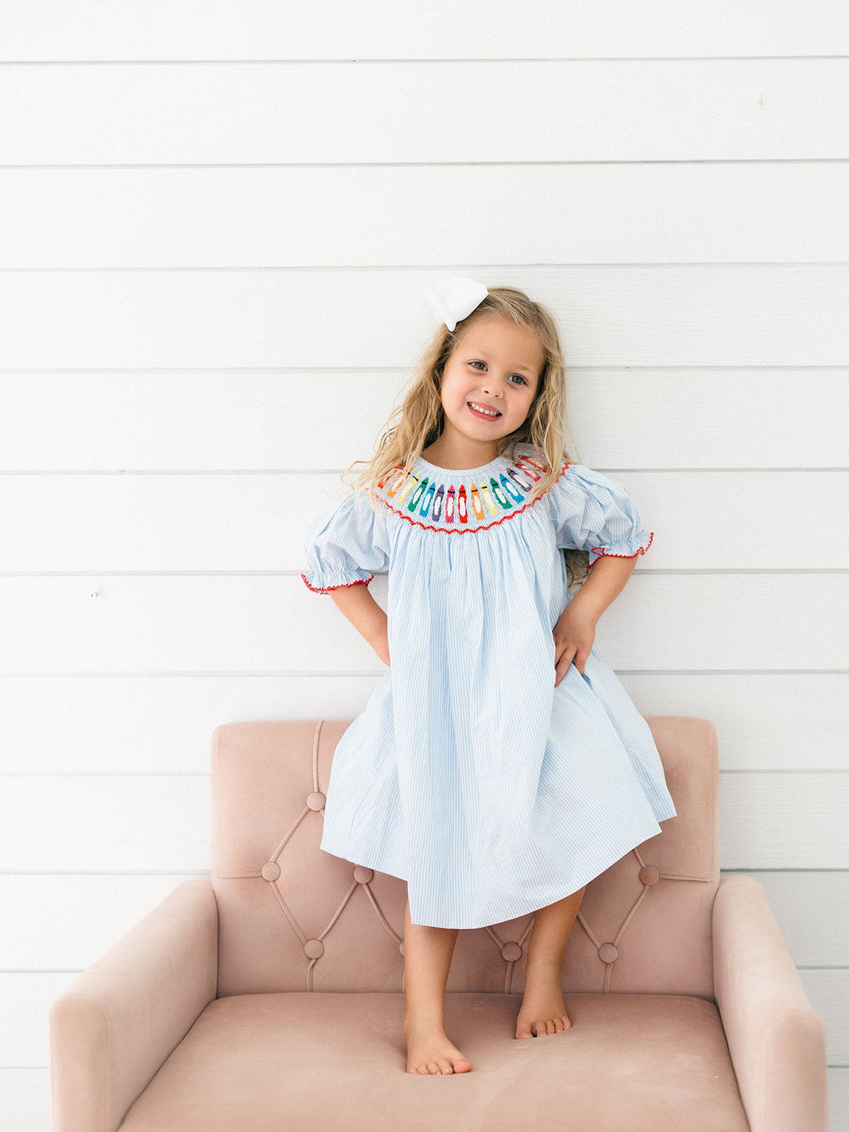 A young girl with blonde hair wearing a light blue smocked dress with colorful crayon embroidered details around the neckline, standing barefoot on a dusty pink tufted armchair against a white shiplap wall.