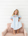 A young girl with blonde hair wearing a light blue smocked dress with colorful crayon embroidered details around the neckline, standing barefoot on a dusty pink tufted armchair against a white shiplap wall.