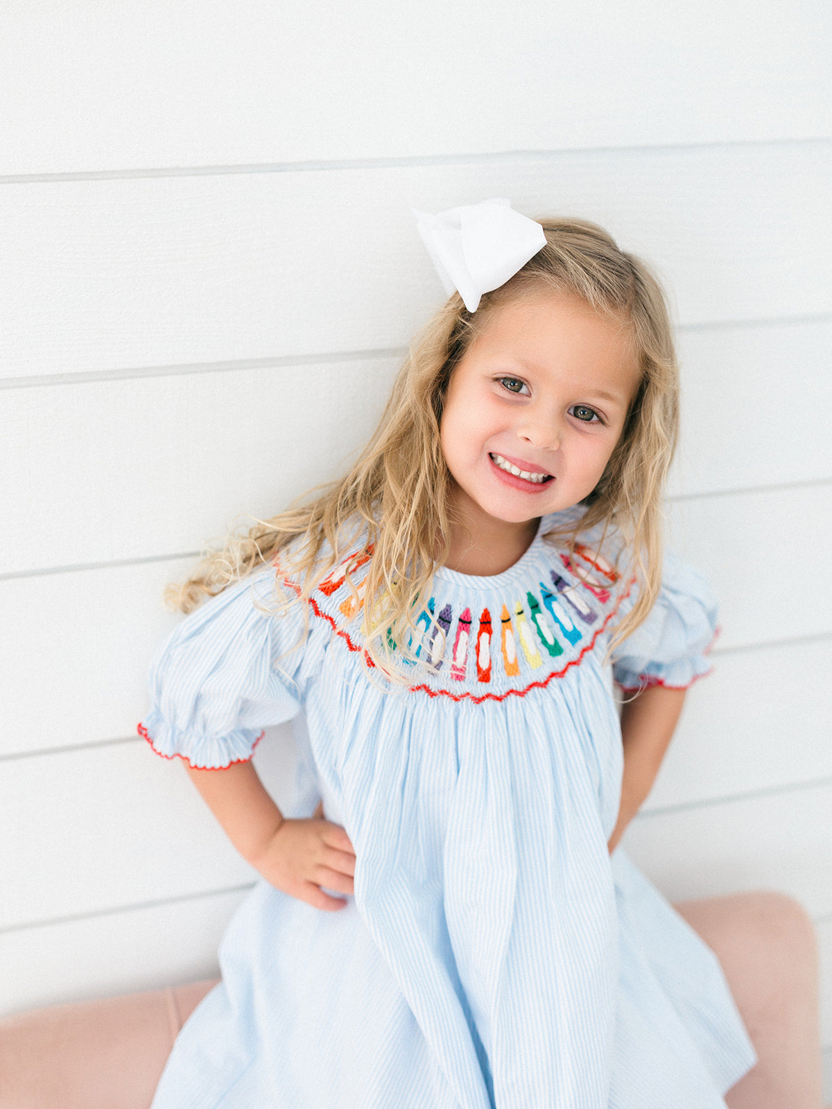 A young girl with blonde hair wearing a light blue smocked dress with colorful crayon embroidered neck detailing and a white bow, posing against a white paneled wall.