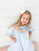 A young girl with blonde hair wearing a light blue smocked dress with colorful crayon embroidered neck detailing and a white bow, posing against a white paneled wall.