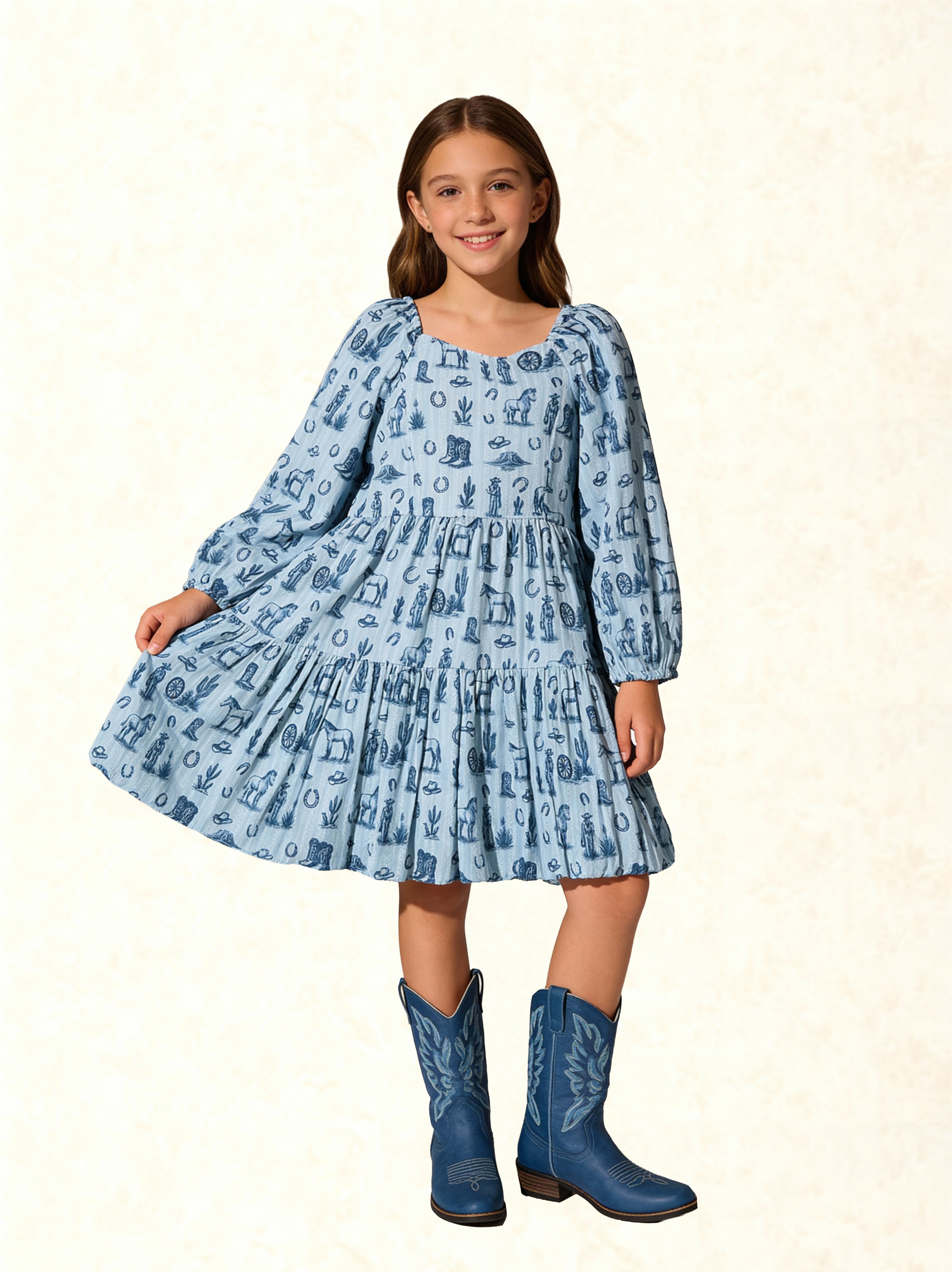 A young girl modeling a light blue long-sleeve tiered dress with a dark blue western-themed print, paired with matching blue cowboy boots, set against a light beige textured background.