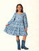 A young girl modeling a light blue long-sleeve tiered dress with a dark blue western-themed print, paired with matching blue cowboy boots, set against a light beige textured background.