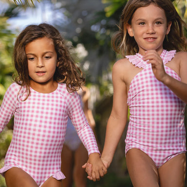 Two young girls wearing pink gingham swimsuits holding hands outdoors.
