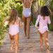 Two children in pink checkered swimsuits running on a wooden path with a woman in a blue swimsuit following them.