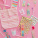 A flat-lay arrangement of pastel pink stationery and tote bag accessories on a pink background, including a large pink tote bag with lined guest writing fields, colorful sticky notes, pens, clear cups, glittery bookmarks, and patterned paper strips.
