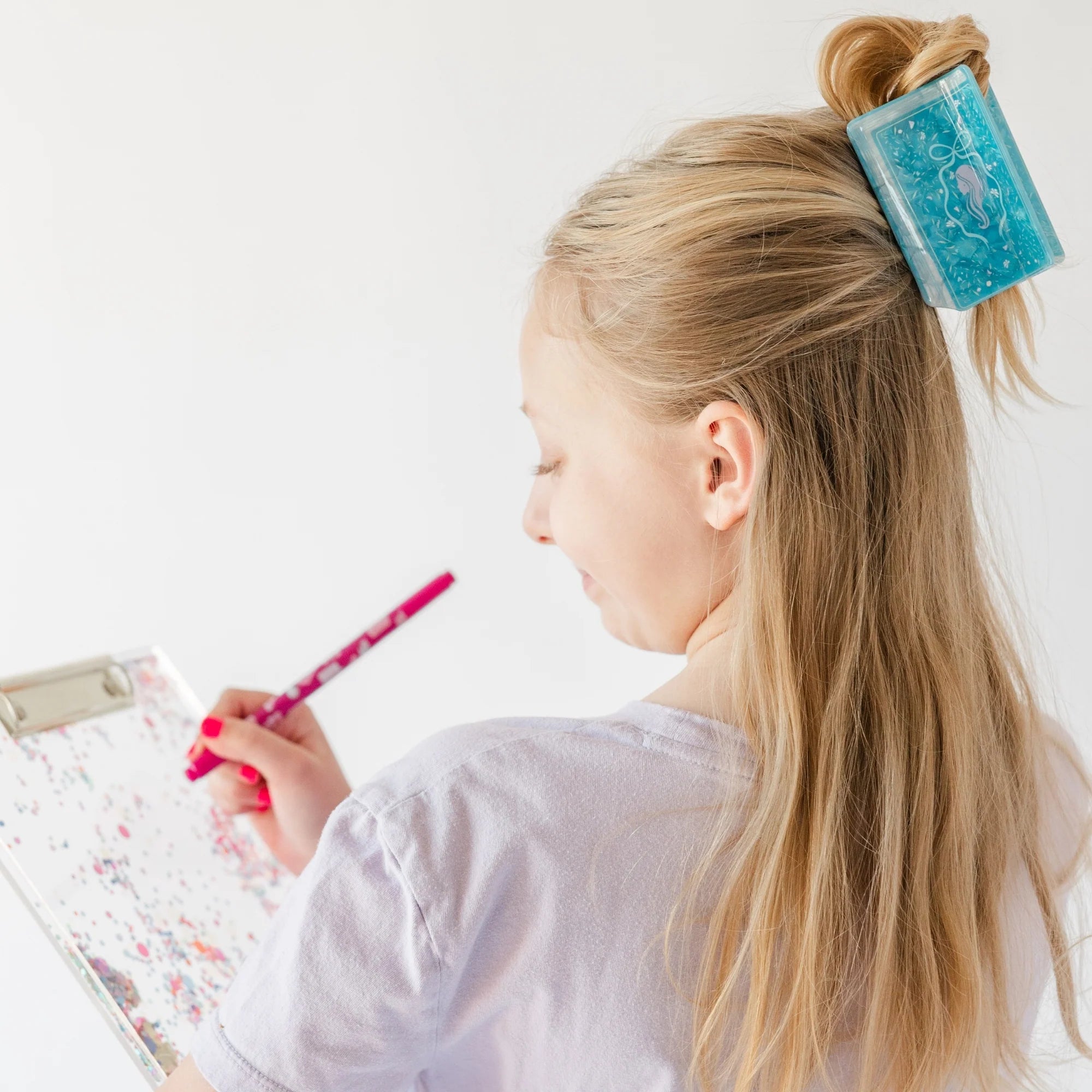 Young girl with a blue hair clip holding a pink pen, sitting at a table with a white background