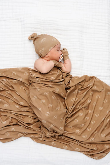 A newborn baby lying on a white textured blanket, wrapped in a brown swaddle blanket with a repeating football pattern, wearing a matching brown knit hat and mittens.