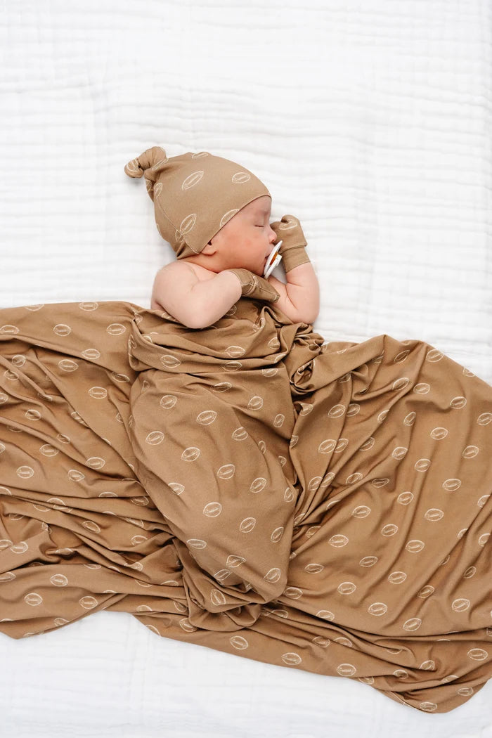 A newborn baby lying on a white textured blanket, wrapped in a brown swaddle blanket with a repeating football pattern, wearing a matching brown knit hat and mittens.