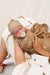 A sleeping newborn baby swaddled in a brown swaddle blanket with a matching hat, resting on a mother's lap. The swaddle and hat feature a light beige football pattern.